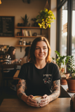 Woman holding a coffee cup in a cozy cafe setting wearing a seven swords tattoo shirt
